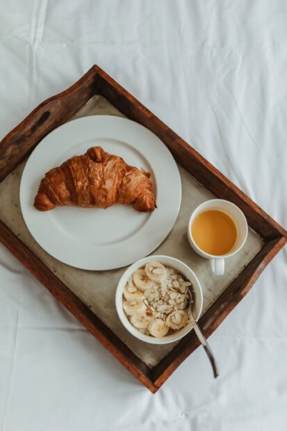 A fresh croissant on a white plate with a bowl of banana and almond slices on a tray