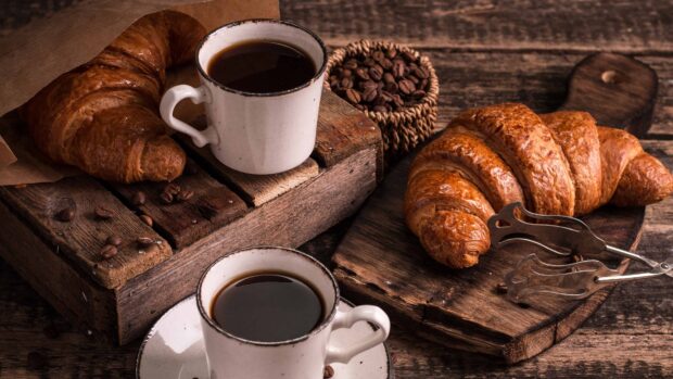 Fresh croissant with coffee cups on rustic wooden table surrounded by coffee beans