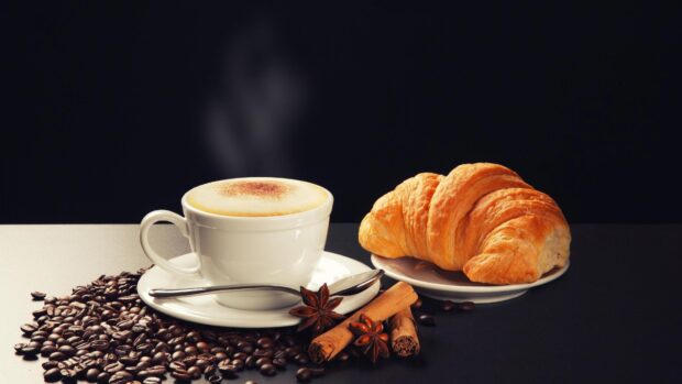 A croissant next to a cup of coffee surrounded by coffee beans and spices