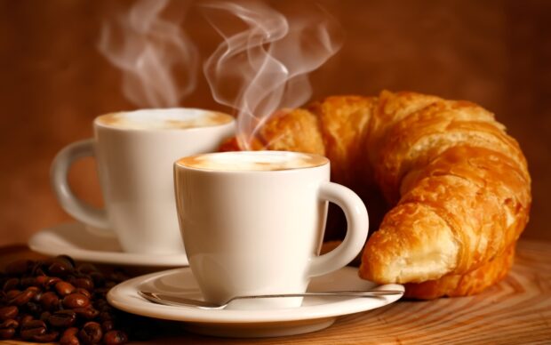 A fresh croissant with steaming coffee cups and coffee beans on a wooden surface