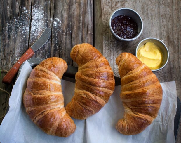 Three fresh croissant breads on a wooden table with jam and butter spreads