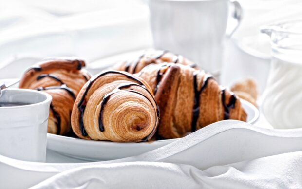 Fresh croissant with chocolate drizzle on a white plate alongside a cup of coffee