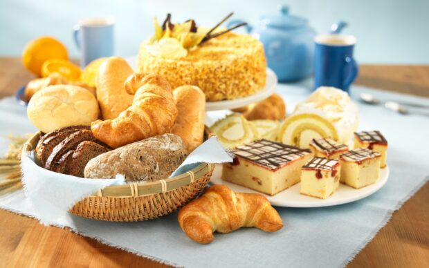 A fresh croissant among various bread and desserts on a table with coffee cups