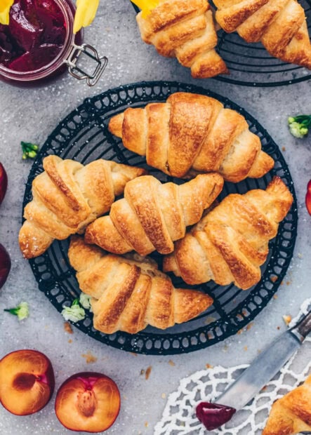 Freshly baked croissant surrounded by plums and jam on a table with flowers