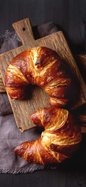 Two golden croissant breads placed on a wooden board and dark fabric background