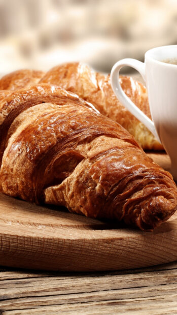 A flaky croissant placed on a wooden board next to a white coffee cup