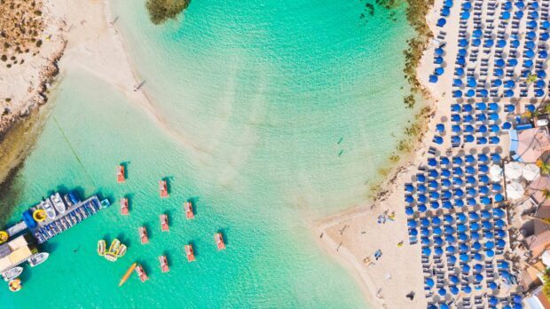 Aerial view of Cyprus turquoise sea with boats and organized blue umbrellas on sandy beach
