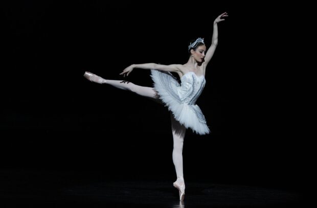 A ballet dancer performing an arabesque pose on stage wearing a white tutu and tiara
