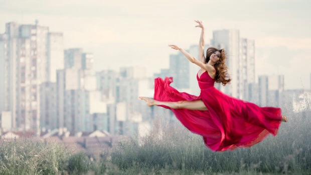 A graceful dancer in a flowing red dress performing a leap outdoors in front of city buildings