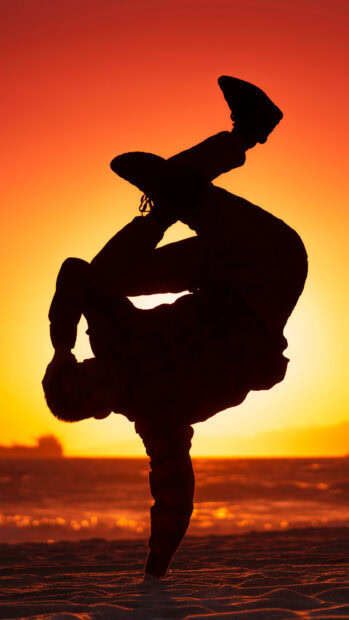 A dancer performing a breakdance pose on one hand during a sunset on the beach