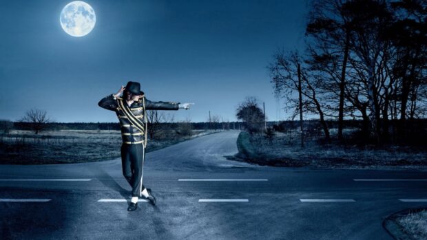 A dancer dressed in a black outfit and hat performing a pose on a nighttime road under a full moon