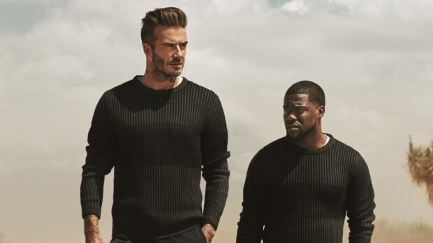 David Beckham standing outdoors wearing a black sweater next to another man with a cloudy sky background