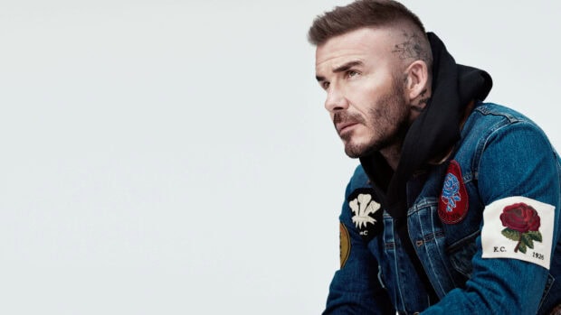 David Beckham wearing a decorated denim jacket and hoodie with visible tattoos on a light background