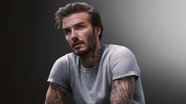 David Beckham with tattoos and beard posing in a grey t shirt with a serious expression