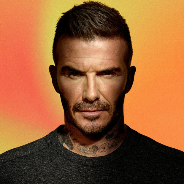 David Beckham with beard and neck tattoos posing against a vibrant orange background