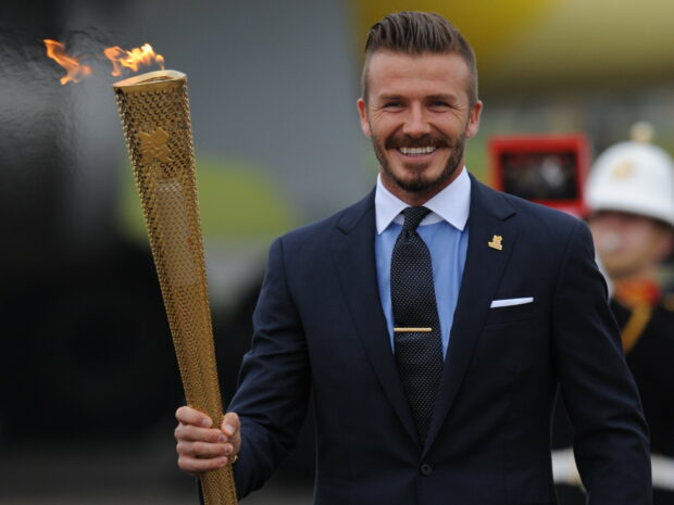 David Beckham holding the Olympic torch and smiling in a dark suit and tie