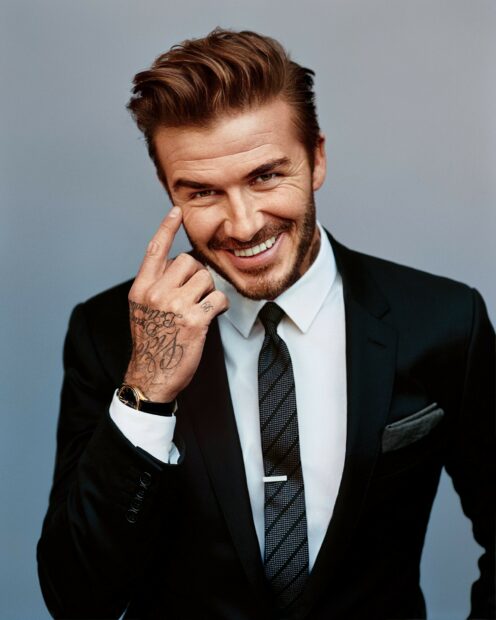 David Beckham smiling confidently in a black suit and tie with visible tattoos on his hand