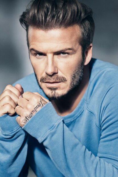 David Beckham wearing a blue sweater with a focused expression and visible tattoo on his hand