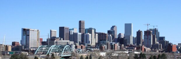 The Denver skyline features tall buildings and a clear blue sky with a light blue arched bridge in the foreground