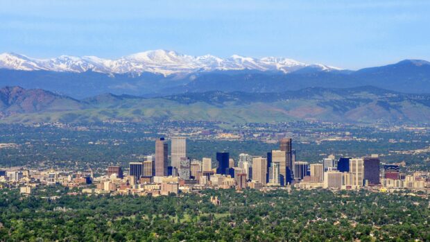 The Denver skyline features tall buildings with mountain ranges in the background under a clear blue sky