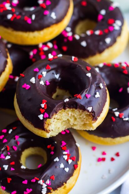 A close up of donut topped with chocolate and heart shaped sprinkles with a bite taken out