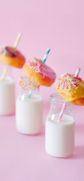Colorful donut with sprinkles on a straw above a glass of milk on a pink background