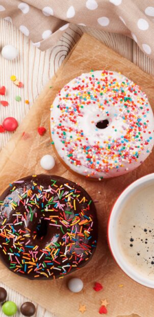 Two donuts with colorful sprinkles and a cup of coffee on a wooden table