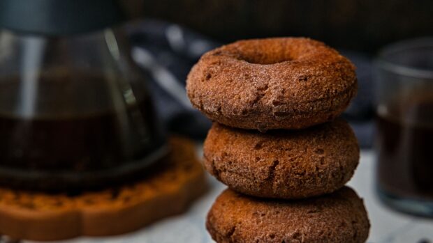 Three stacked chocolate donuts with a rich texture and a cup of coffee in the background