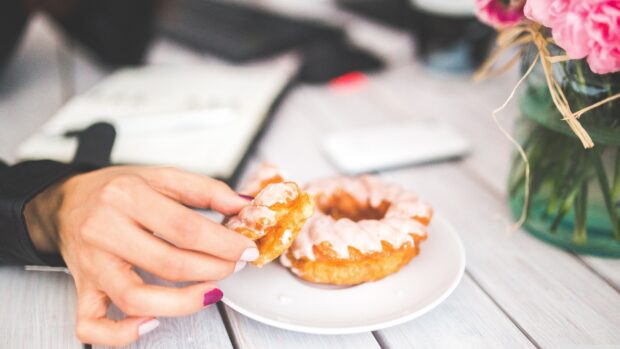 A hand holding a glazed donut with a plate of donuts on a wooden table