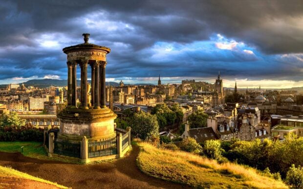 Historical monument surrounded by greenery overlooking the Edinburgh skyline at sunset with dramatic clouds