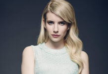 Emma Roberts wearing a light blue sleeveless top posing confidently against a dark background