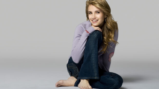 Emma Roberts sitting barefoot in casual jeans and purple sweater smiling at the camera