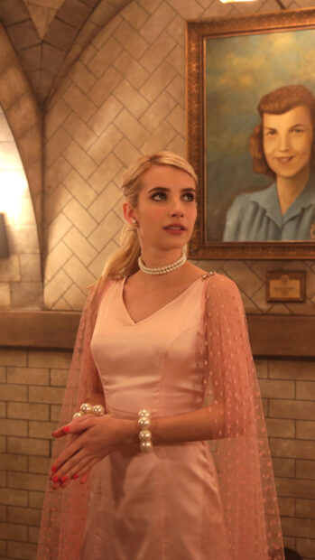Emma Roberts wearing a pink dress and pearl jewelry standing indoors