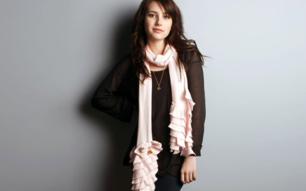 Emma Roberts wearing a black top and pink ruffled scarf standing against a gray background