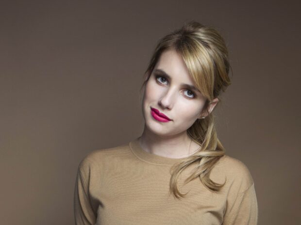 Emma Roberts with blonde hair and red lips wearing a brown sweater in a studio setting