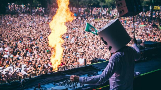 A DJ wearing a helmet performs at a festival with a large crowd and fire effects in the background