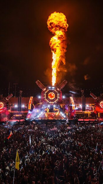 A large festival gathering with pyrotechnics and fire effects lighting up the night sky