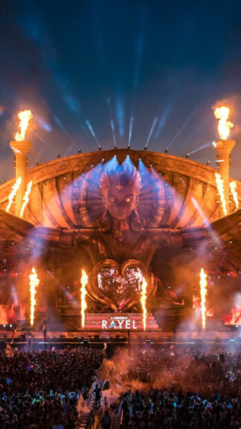 A large festival stage with a glowing statue and fiery pyrotechnics during a night event