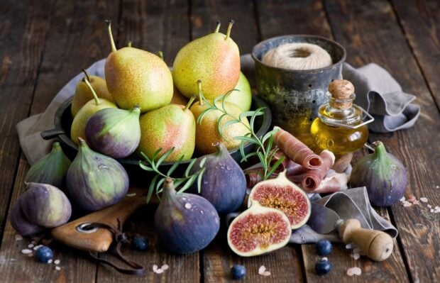 A rustic wooden table with fresh figs and pears arranged alongside herbs and olive oil
