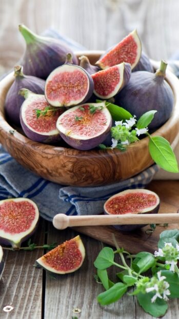 Fresh fig slices and whole figs in a wooden bowl with green leaves on a rustic table