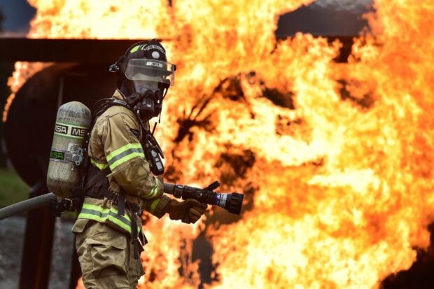 Fireman in full gear spraying water at a large fire blaze with flames in the background