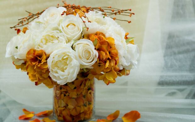 A flower bouquet with white roses and orange flowers in a glass vase filled with petals