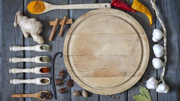 A wooden cutting board surrounded by garlic and various spices on a rustic wooden surface