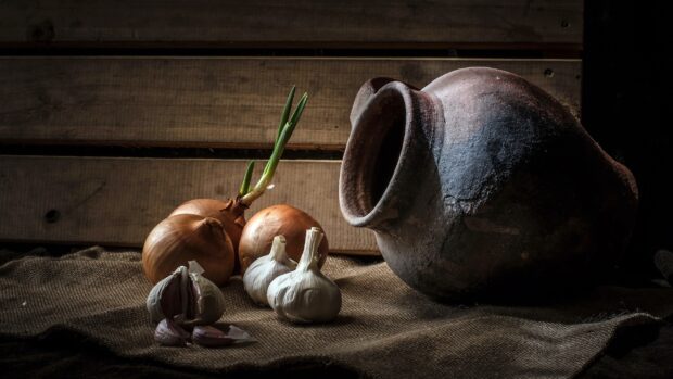 Fresh garlic bulbs placed next to onions and a rustic clay pot on a burlap cloth