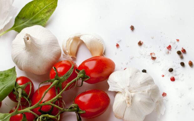 Fresh garlic cloves surrounded by ripe cherry tomatoes and basil leaves on a white surface