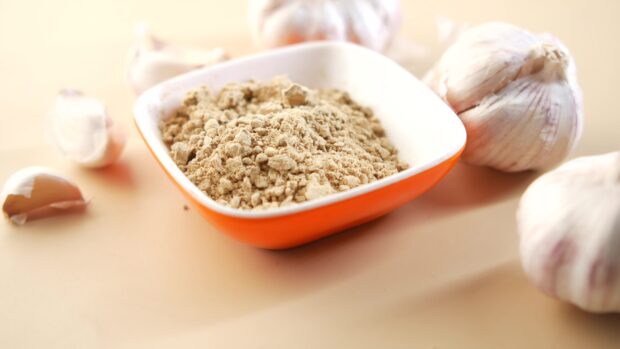 Ground garlic in a bowl surrounded by whole garlic bulbs on a beige surface