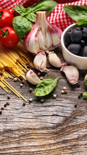 Fresh garlic cloves and a garlic bulb surrounded by basil leaves and spices on wooden surface