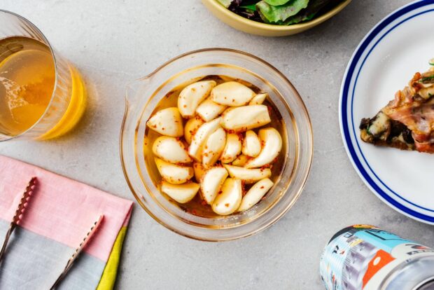 A bowl of peeled garlic cloves marinated with chili flakes in oil on a kitchen countertop