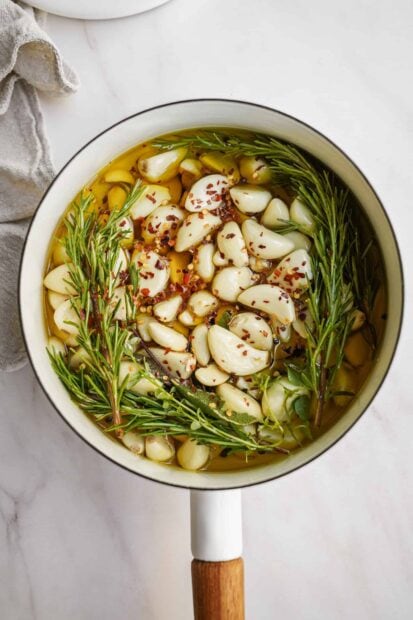 Fresh garlic cloves simmering in olive oil with herbs and spices in a white pot