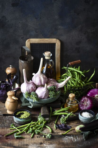 Fresh garlic bulbs surrounded by fresh herbs and green vegetables on a rustic wooden table